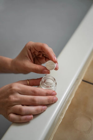 A hand reaches for a small glass jar of MADISON AND GREEN - REST YOUR EYES IN THE BATH by MADISON GREEN, filled with soothing white cream, sitting on the edge of a bathtub beside beige tiles—creating an ideal setting for relaxing eye therapy or pairing your soak with essential oil tablets for the ultimate spa-like experience.