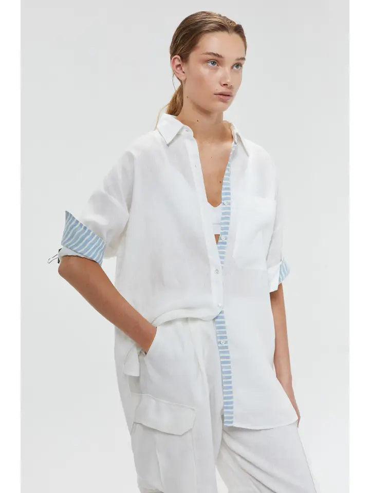 A person stands against a plain background, wearing the LANHTROPY - ISLAND LINEN OVERSIZED SHIRT from LANHTROPY, crafted from premium organic linen. The shirt features blue and white striped detailing on the cuffs and inner placket, paired with white pants. Elegant shell buttons accentuate the shirt. Their hands rest in the pockets of their pants as they gaze directly at the camera.
