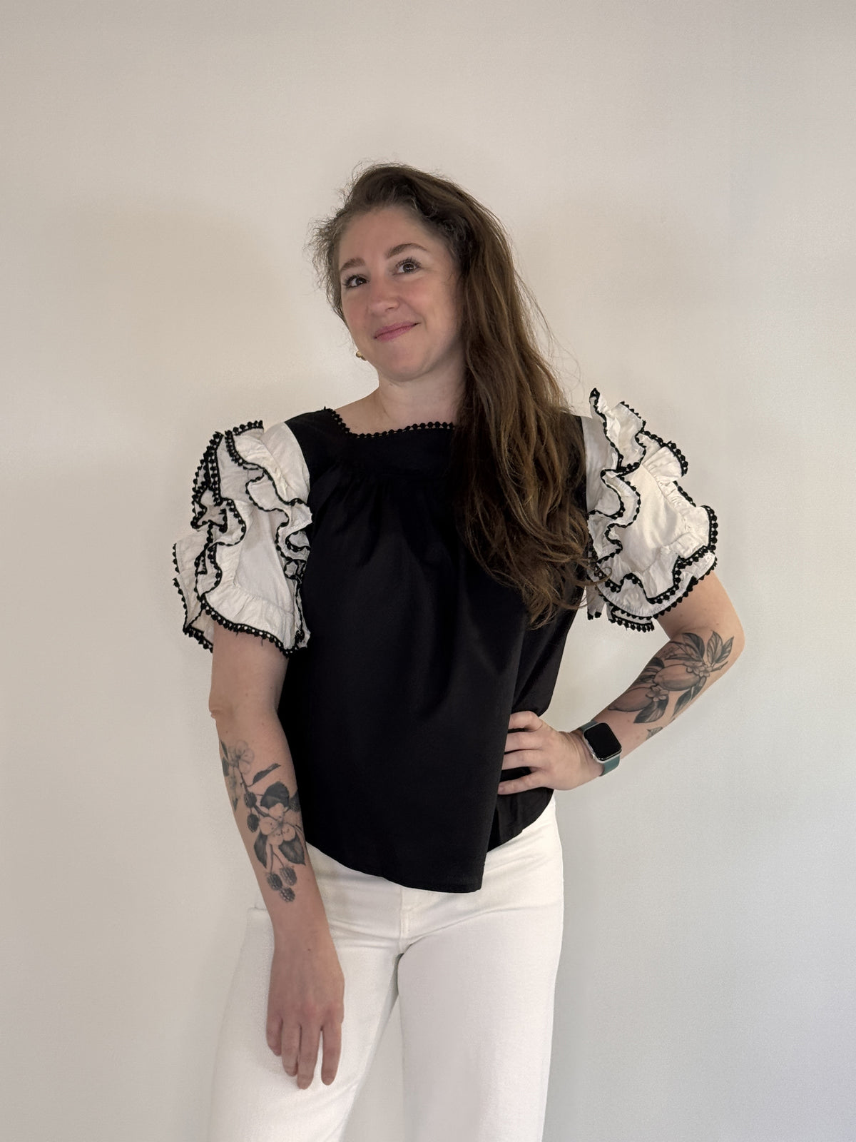 A person with long brown hair and tattoos on both arms stands against a plain background, smiling. They wear the FARYLINA - DAHLIA TOP by FARYLINA—a black and white statement blouse with ruffled sleeves—white pants, and an Apple Watch.