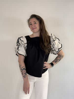A person with long brown hair and tattoos on both arms stands against a plain background, smiling. They wear the FARYLINA - DAHLIA TOP by FARYLINA—a black and white statement blouse with ruffled sleeves—white pants, and an Apple Watch.