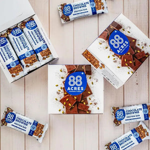 An 88 ACRES - DARK CHOCOLATE BROWNIE HIGH PROTEIN BAR sits on a white wooden surface. The nut-free, vegan, and gluten-free packaging highlights it contains 12g of protein.