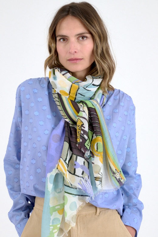 A woman in a light blue textured blouse and beige pants stands with one hand in her pocket, wearing the colorful STORIATIPIC - HEIDI SCARF from STORIATIPIC, its chic cotton fabric billowing stylishly around her neck.