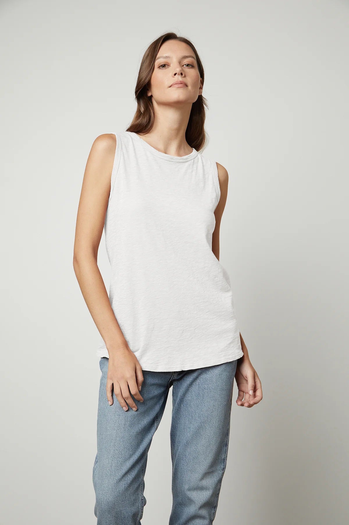 A person with long, brown hair is wearing the VELVET - TAURUS TANK IN COTTON SLUB by VELVET BY GRAHAM &amp; SPENCER and blue jeans while facing the camera. Their right hand is resting on their thigh, and their left hand is visible by their side. The plain, light-colored background complements the tomboy-inspired look.