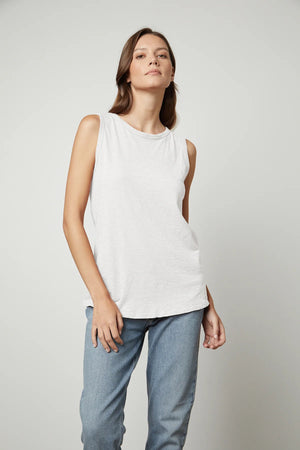 A person with long, brown hair is wearing the VELVET - TAURUS TANK IN COTTON SLUB by VELVET BY GRAHAM & SPENCER and blue jeans while facing the camera. Their right hand is resting on their thigh, and their left hand is visible by their side. The plain, light-colored background complements the tomboy-inspired look.