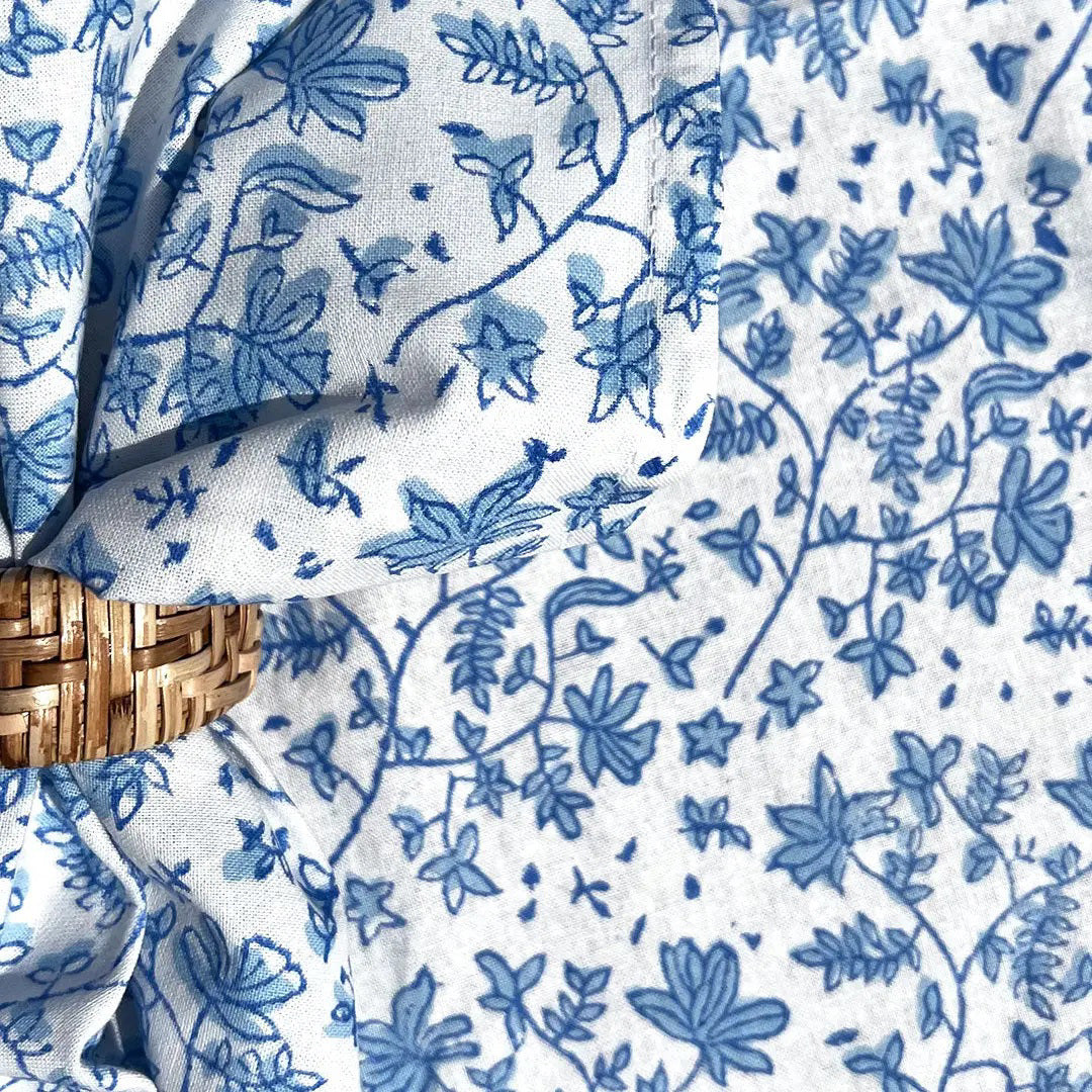 The CHRISTINA DICKSON HOME - WHIMSY FLORAL COTTON NAPKIN, an artisan-crafted blue and white floral fabric made in India, is tied in a knot over a circular rattan mat. Its delicate leaf and flower design complements the mat&#39;s textured pattern.