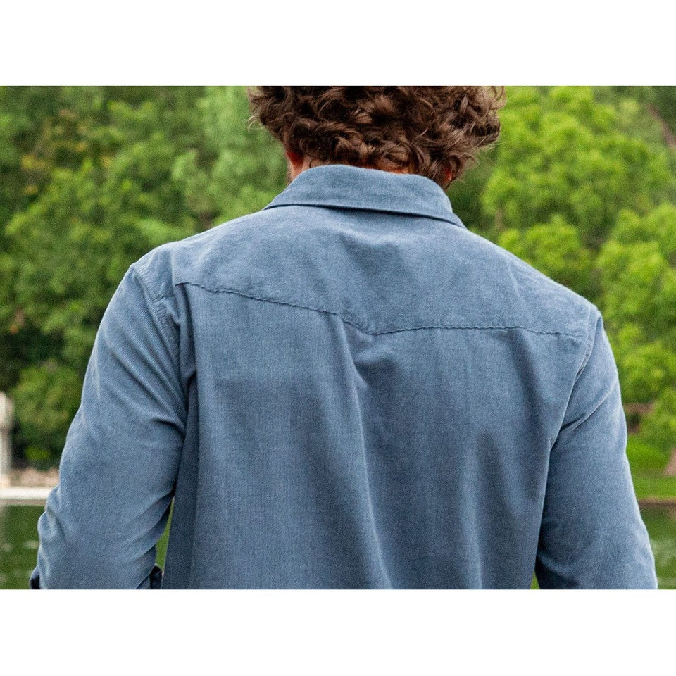 A man is wearing the CRIQUET Long Sleeve Corduroy Pearl Snap Shirt in Slate Blue by CRIQUET, featuring a chest pocket and rolled-up cuffs. He pairs it with brown pants against a white background.