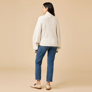 A woman wearing a cream-colored, plush SOFTIES - CABLE KNIT MARSHMALLOW FUNNEL NECK DOUBLE ZIP JACKET with a funnel neck and double zipper, paired with blue jeans.