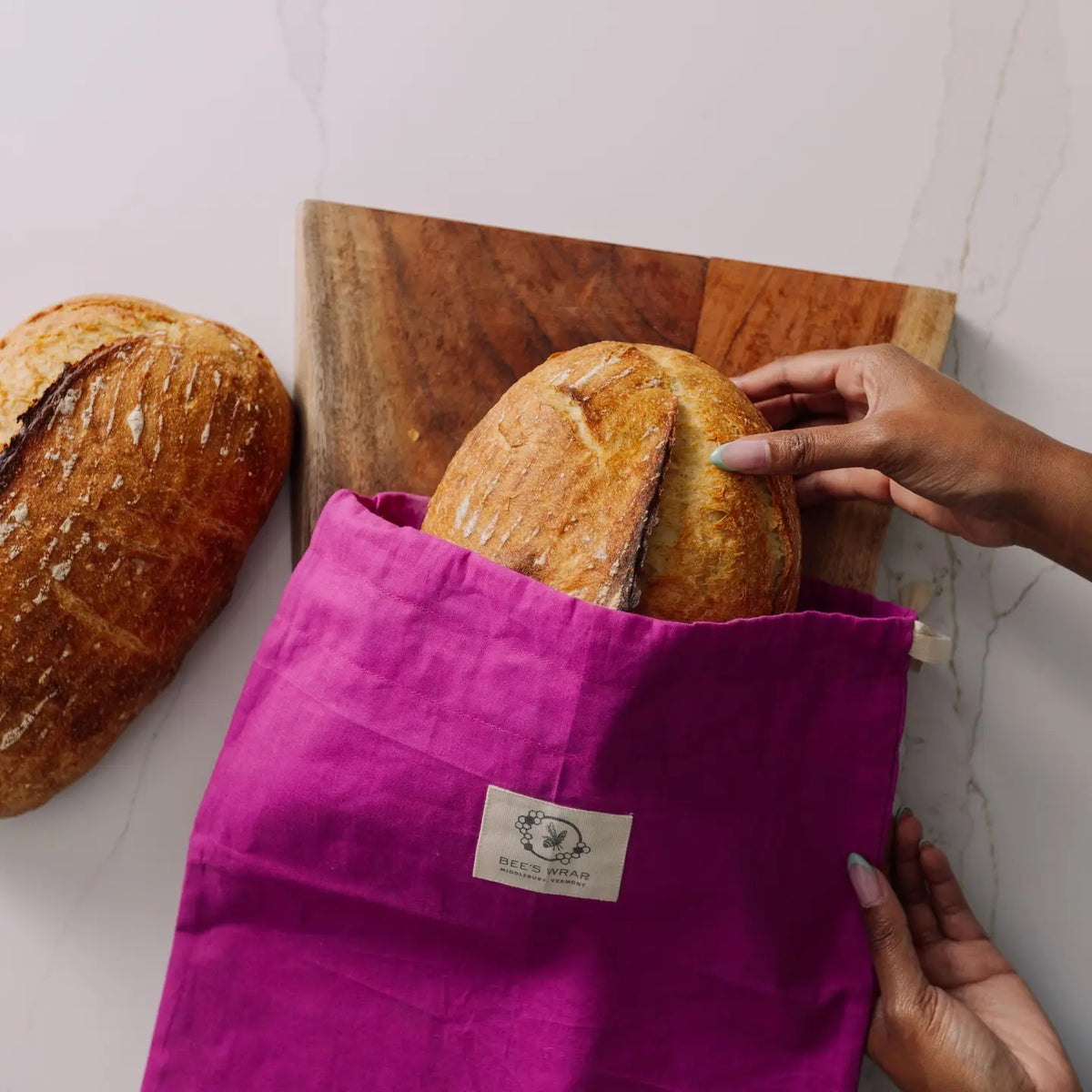 A round loaf of bread in the BEES WRAP - BEESWAX REUSABLE BREAD BAKERY BAG MAGENTA by BEES WRAP, featuring a magenta cloth and floral trim, is placed on a textured tablecloth beside a wooden cutting board, wheat stalks, and a folded napkin.
