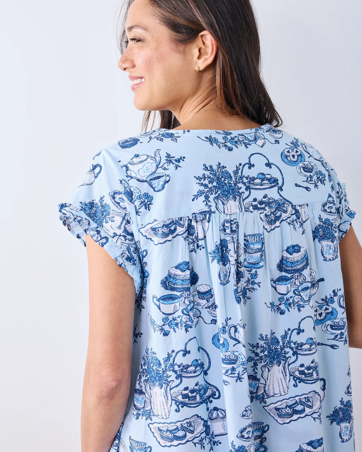 A woman stands barefoot on a wood floor, wearing the PRINTFRESH High Tea Toile Pintuck Nightgown in Notting Hill Blue—a vintage-inspired piece with short sleeves, a ruffled hem, and a charming blue illustrated print. She poses against a white wall with a relaxed smile.