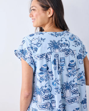 A woman stands barefoot on a wood floor, wearing the PRINTFRESH High Tea Toile Pintuck Nightgown in Notting Hill Blue—a vintage-inspired piece with short sleeves, a ruffled hem, and a charming blue illustrated print. She poses against a white wall with a relaxed smile.