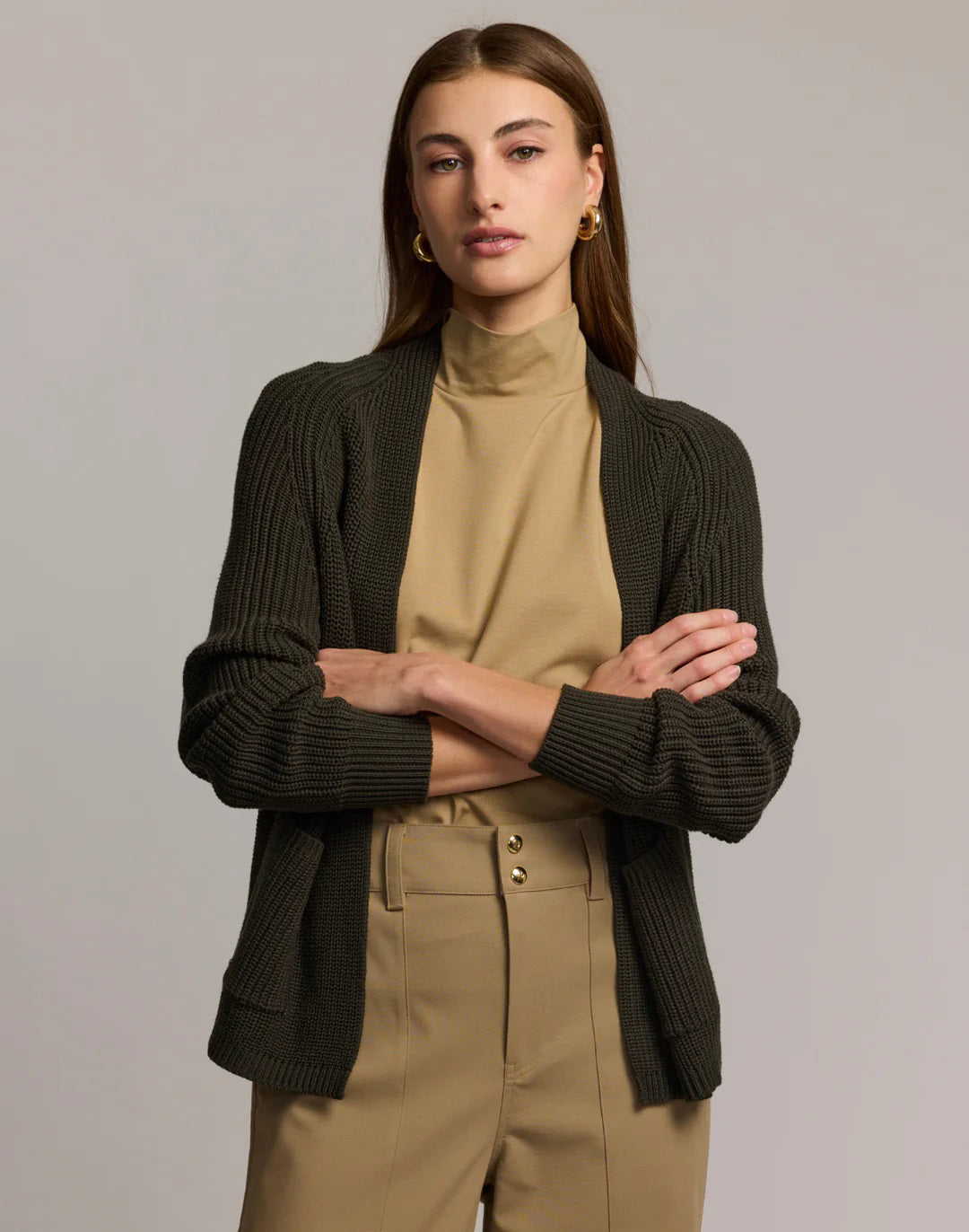 A woman with straight brown hair, in a tan turtleneck and high-waisted tan pants with gold buttons, wears the HINSON WU ANGIE Long Sleeve Cardigan in dark olive green, standing arms crossed against a plain light gray background.