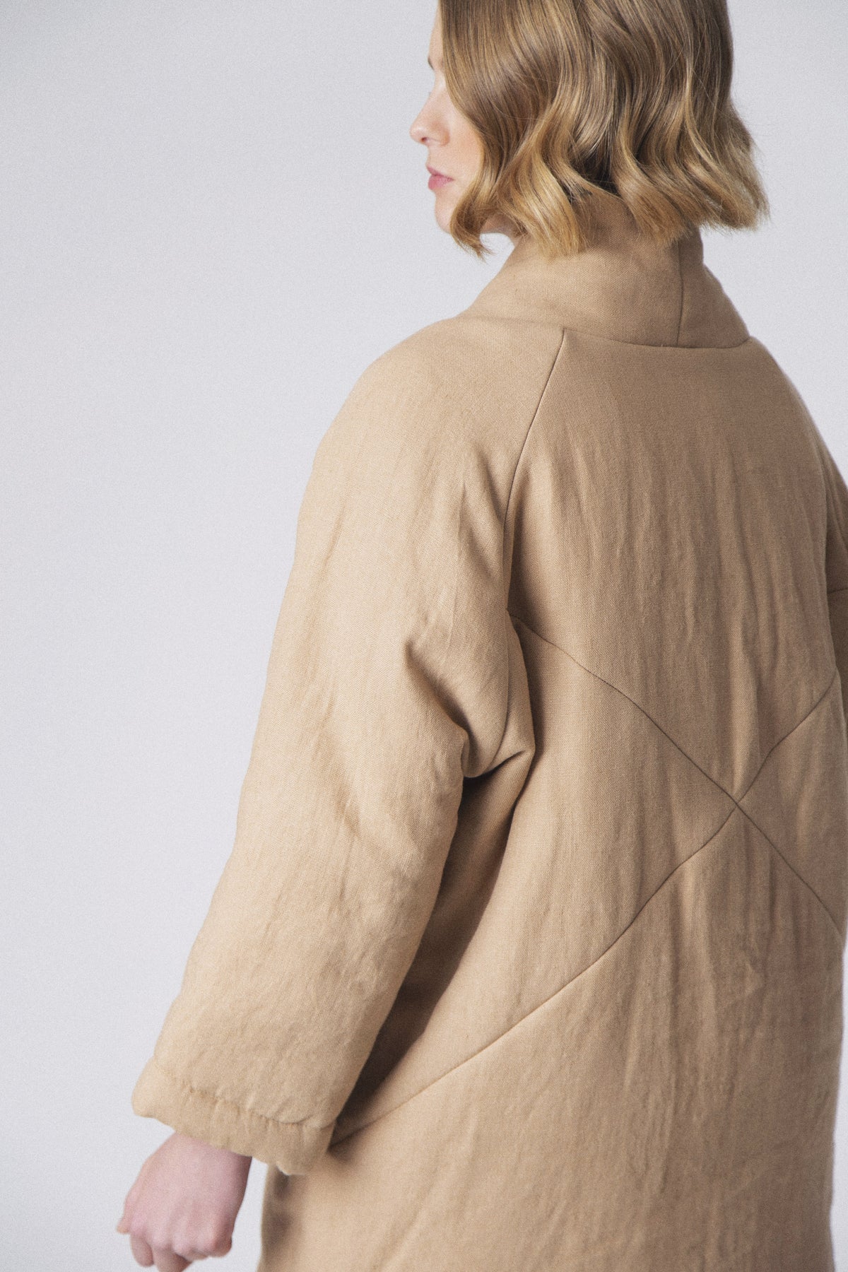 Against a light gray backdrop, a person with short hair models the LOESS MONICA Linen Puffer Coat in beige, complete with matching pants and light slippers. This minimalist outerwear, tied at the waist, captures the essence of transitional weather layering as they stand relaxed.