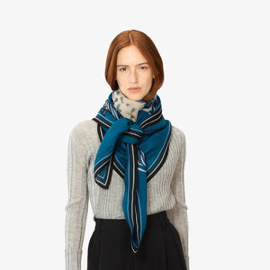 A person with straight, reddish-brown hair models the INOUI EDITIONS - WESTERN SQUARE SCARF by INOUI, a blue and white patterned wool accessory made in India, draped over a light gray ribbed sweater and black pants, standing against a plain white background.