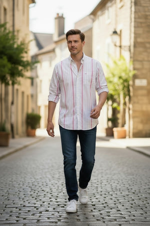 A man in a FRANK & EILEEN FINBAR men's button-up shirt in hot pink and blue stripe, paired with jeans, walks outdoors along a sunlit street lined with buildings and greenery.
