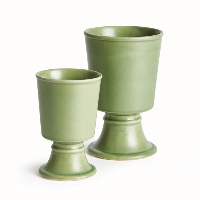 Two green NAPA HOME AND GARDEN - KELCY FOOTED CACHEPOT planters sit on a beige table runner atop a wooden table. The taller, reactive-glaze planter holds a leafy plant, while the shorter one is empty. A white lattice forms the background.