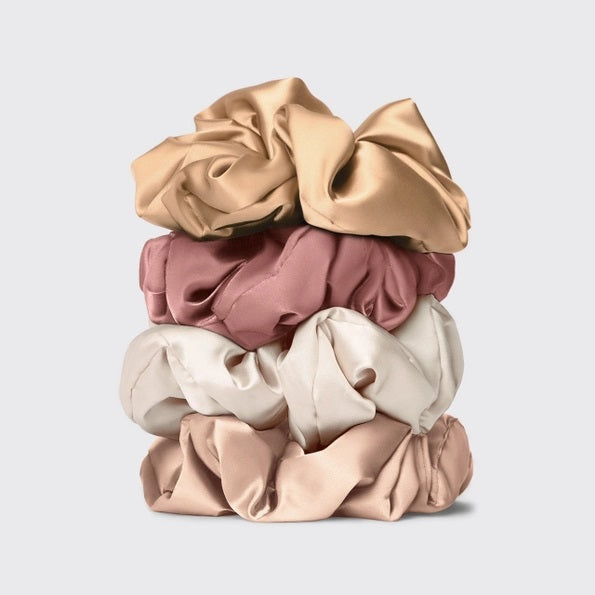 A package of KITSCH PILLOW SATIN SCRUNCHIES IN BLUSH sits behind four stacked scrunchies in beige, pink, cream, and champagne shades. The packaging displays a woman’s face and reads “Pillow Scrunchies in Satin” by KITSCH, showcasing this gentle, damage-free hair accessory ideal for soft styling.