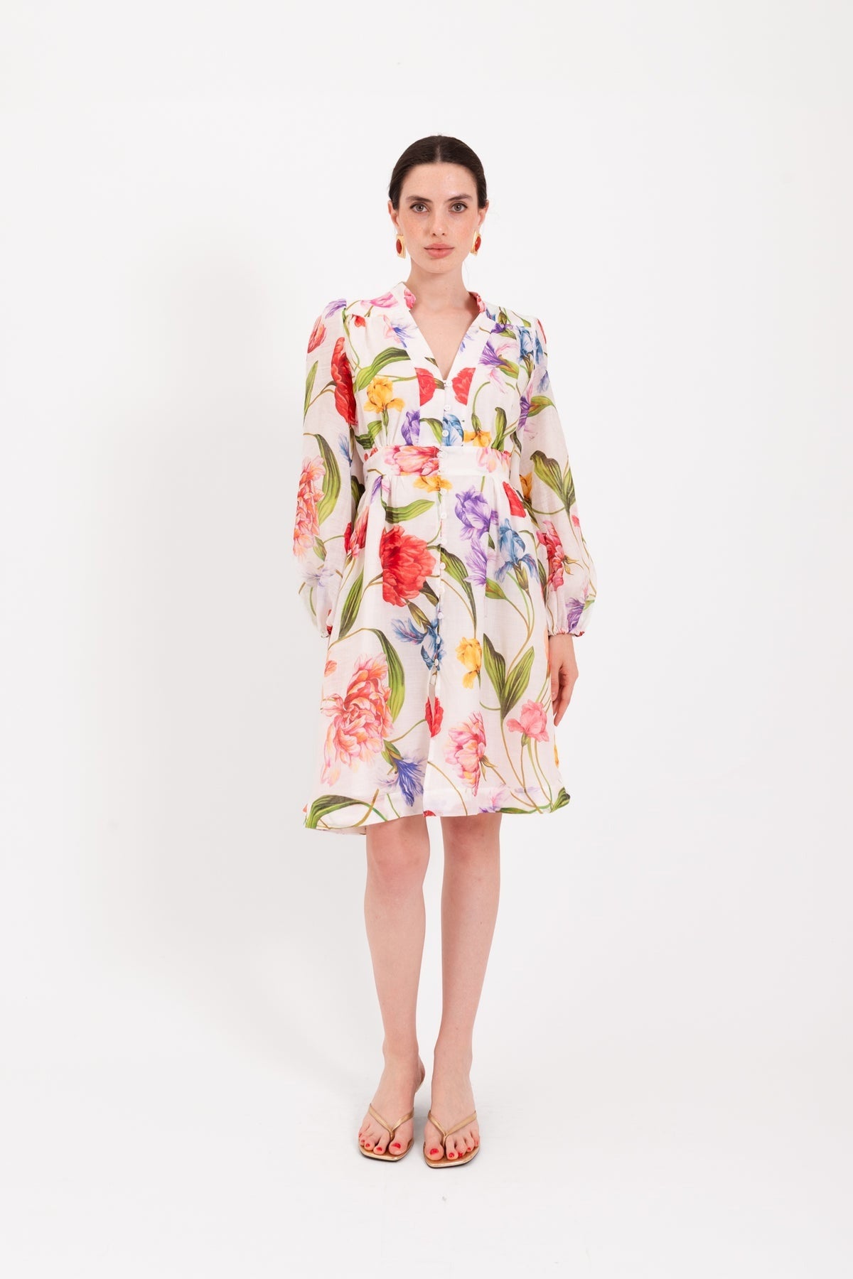 A woman stands against a white background, wearing the LAVI - CETERA FLORAL DRESS, a knee-length, long-sleeve dress by LAVI featuring a vibrant botanical print. She pairs it with open-toed sandals, her hair pulled back and her expression neutral.