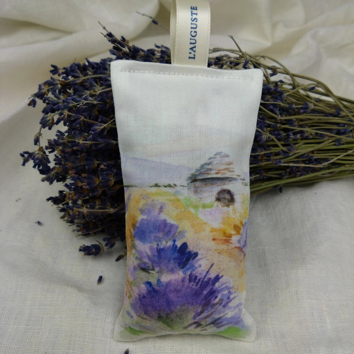 A L'AUGUSTE PROVENCE - FRENCH LAVENDER SACHET FROM PROVENCE BORIE, featuring a watercolor-style lavender field and house, rests in front of dried lavender on a light cloth background.