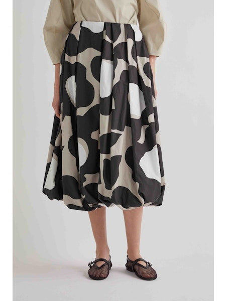 A woman stands against a plain background wearing a beige long-sleeve top, black sandals, and the MEIMEIJ BLACK MOONBEAM SKIRT by MEIMEIJ—a voluminous midi skirt featuring large white and gray abstract shapes.