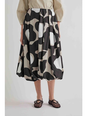 A woman stands against a plain background wearing a beige long-sleeve top, black sandals, and the MEIMEIJ BLACK MOONBEAM SKIRT by MEIMEIJ—a voluminous midi skirt featuring large white and gray abstract shapes.