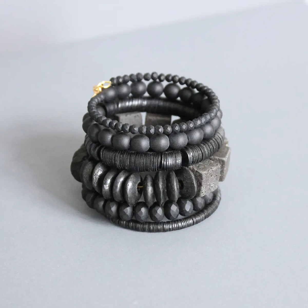 The DAVID AUBREY BLACK WRAP BRACELET, by DAVID AUBREY, features a stack of black beaded strands with round, disc-shaped, faceted, and chunky lava beads displayed on a light gray surface.