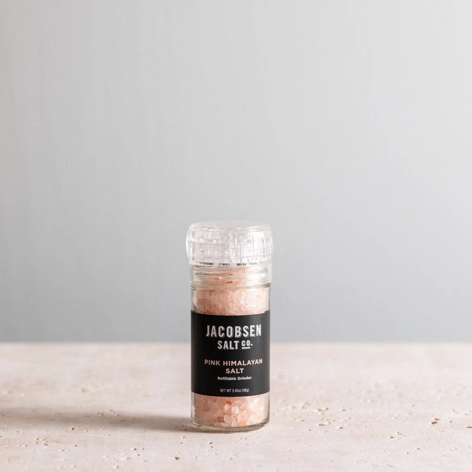 A refillable glass grinder labeled "JACOBSEN SALT - GRINDER PINK HIMALAYAN SALT" from JACOBSEN SALT CO. rests on a light stone surface with a plain gray background, filled with Himalayan pink salt crystals.