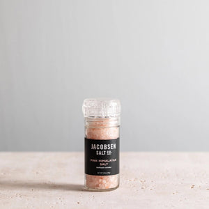 A refillable glass grinder labeled "JACOBSEN SALT - GRINDER PINK HIMALAYAN SALT" from JACOBSEN SALT CO. rests on a light stone surface with a plain gray background, filled with Himalayan pink salt crystals.
