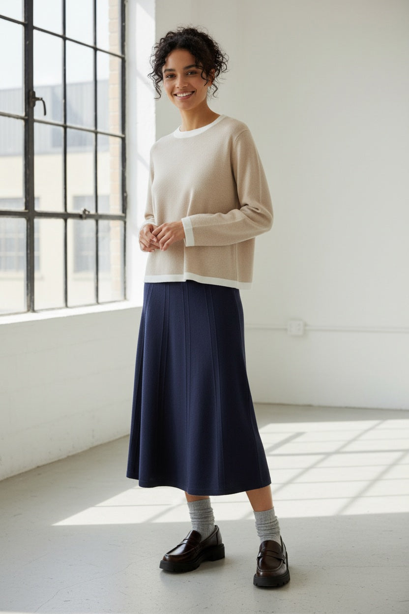 A person, shown from the waist down, wears the ANNECLAIRE knit wool flare skirt in beige with gray socks and black loafers, standing against a light plain background.