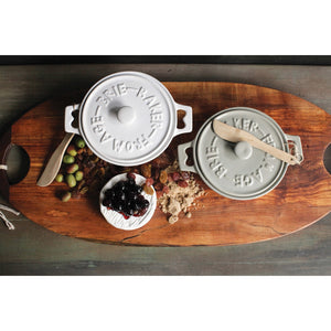 Set of two CREATIVE COOP stoneware brie bakers with lids—one gray, one white—each featuring handles and a matching wooden spreader tied on top.