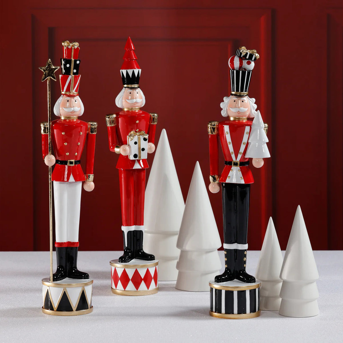 THE TWINKLE TROOP by ZODAX features individual 3.5&quot; x 18&quot; festive nutcracker figurines in red, black, and white, each on a patterned base, displayed among white ceramic cone-shaped trees against a red backdrop.