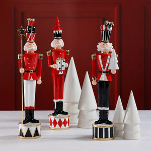 THE TWINKLE TROOP by ZODAX features individual 3.5" x 18" festive nutcracker figurines in red, black, and white, each on a patterned base, displayed among white ceramic cone-shaped trees against a red backdrop.