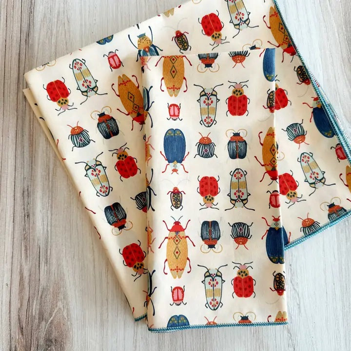 Introducing the DOT AND ARMY - BEETLE DINNER NAPKIN by DOT AND ARMY: a folded white cotton napkin adorned with a vibrant pattern of beetles and bugs in red, blue, yellow, and black hues, finished with blue edge stitching. Perfect as reusable napkins or charming beetle dinner accents for your table setting.