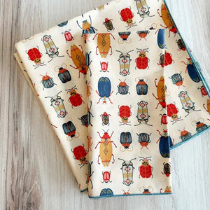 Introducing the DOT AND ARMY - BEETLE DINNER NAPKIN by DOT AND ARMY: a folded white cotton napkin adorned with a vibrant pattern of beetles and bugs in red, blue, yellow, and black hues, finished with blue edge stitching. Perfect as reusable napkins or charming beetle dinner accents for your table setting.
