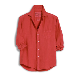 A woman in sunglasses and white shorts smiles outdoors, wearing the FRANK & EILEEN EILEEN Relaxed Button Up Shirt in Washed Linen Summer Red. She stands by a wooden table and chairs, with a house featuring large windows behind her.