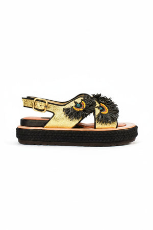 Discover the MEHER KAKALIA - RIO BLOC SANDAL IN CHARM GOLD, featuring a gold glitter design with a wide black sole, vibrant fringe in black, red, blue, and yellow, an open toe, ankle strap with buckle, and a thick textured base for a striking statement.