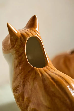 The QUAIL CERAMICS - SQUASH CAT FLOWER VASE by QUAIL DESIGNS is a hand-painted stoneware vase shaped like a sitting orange cat, with green foliage and yellow flowers appearing to grow from its back.