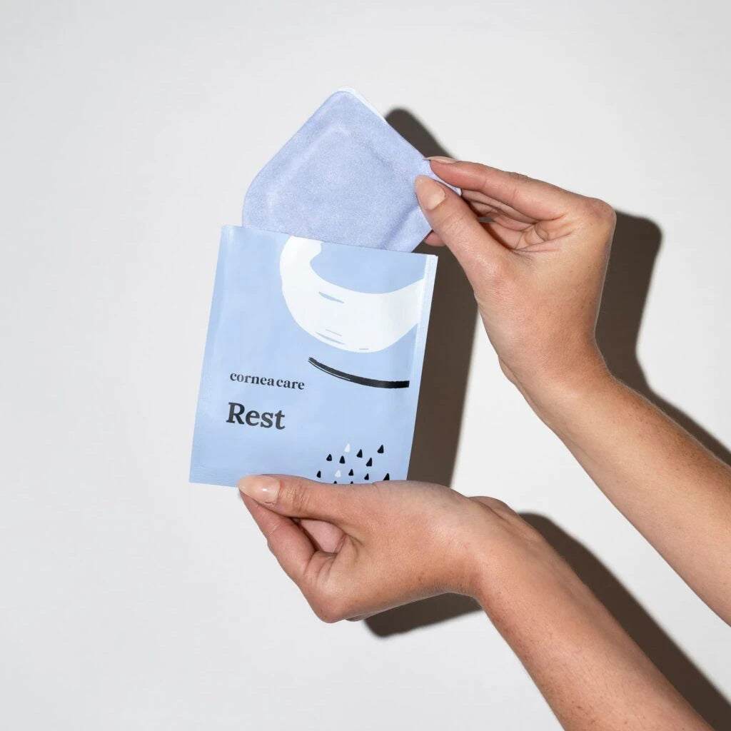 A blue and white CORNEACARE - REST SELF HEATING WARM COMPRESSES package with a crescent illustration lies next to an unfolded blue eye mask with white straps, designed by CORNEA CARE to soothe eye dryness and provide skincare benefits.