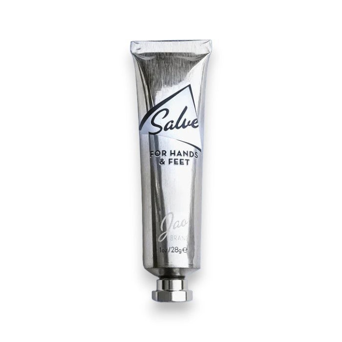 A silver tube labeled "JAO BRAND - SALVE FOR HANDS AND FEET" by JAO LIMITED, featuring essential oils, is placed inside a small white bowl. Beside the tube is a metallic golden key-like tool. The bowl sits on a marble surface.