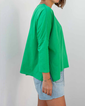 A woman with long, straight hair stands against a white wall, wearing the MERSEA Catalina Slub Tee in blue paired with white pants. The midweight textured cotton tee is from MERSEA. She looks at the camera with a neutral expression.