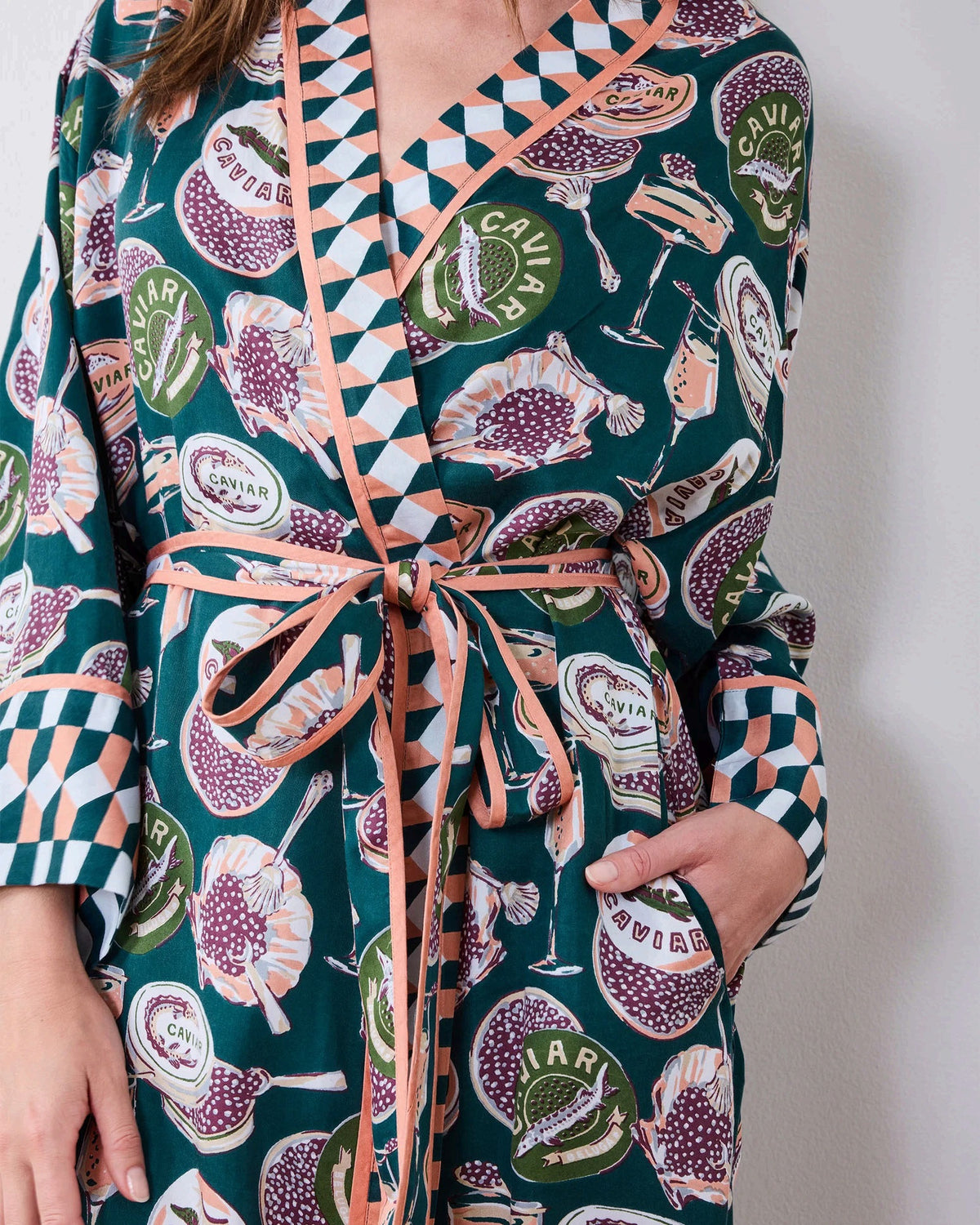 A woman stands barefoot against a white wall, smiling and holding a white cup in one hand as she poses slightly angled to the side. She is wearing the PRINTFRESH Champagne & Caviar Eco Satin Robe in Peacock Feather—a long, colorful eco-friendly robe featuring a vibrant botanical print with peacock feathers and checkered trim.