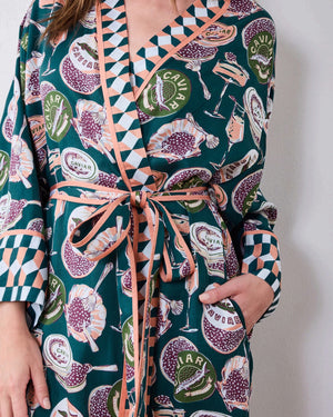 A woman stands barefoot against a white wall, smiling and holding a white cup in one hand as she poses slightly angled to the side. She is wearing the PRINTFRESH Champagne & Caviar Eco Satin Robe in Peacock Feather—a long, colorful eco-friendly robe featuring a vibrant botanical print with peacock feathers and checkered trim.