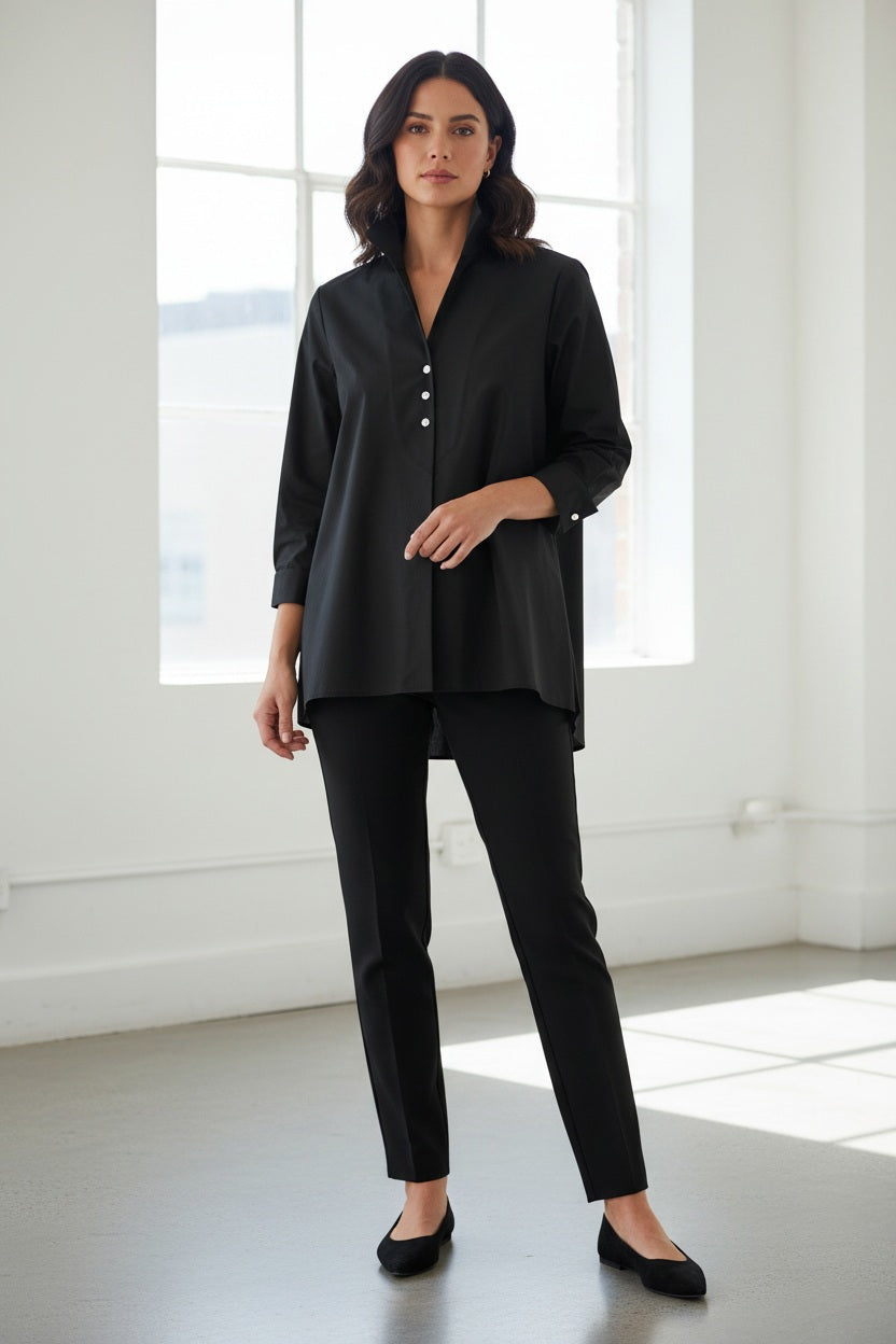 A woman with straight brown hair models the HINSON WU BETTY LONG SLEEVE TUNIC, a loose-fitting, white, A-line button-up shirt with a standing collar and rolled sleeves, paired with black pants against a plain light background.