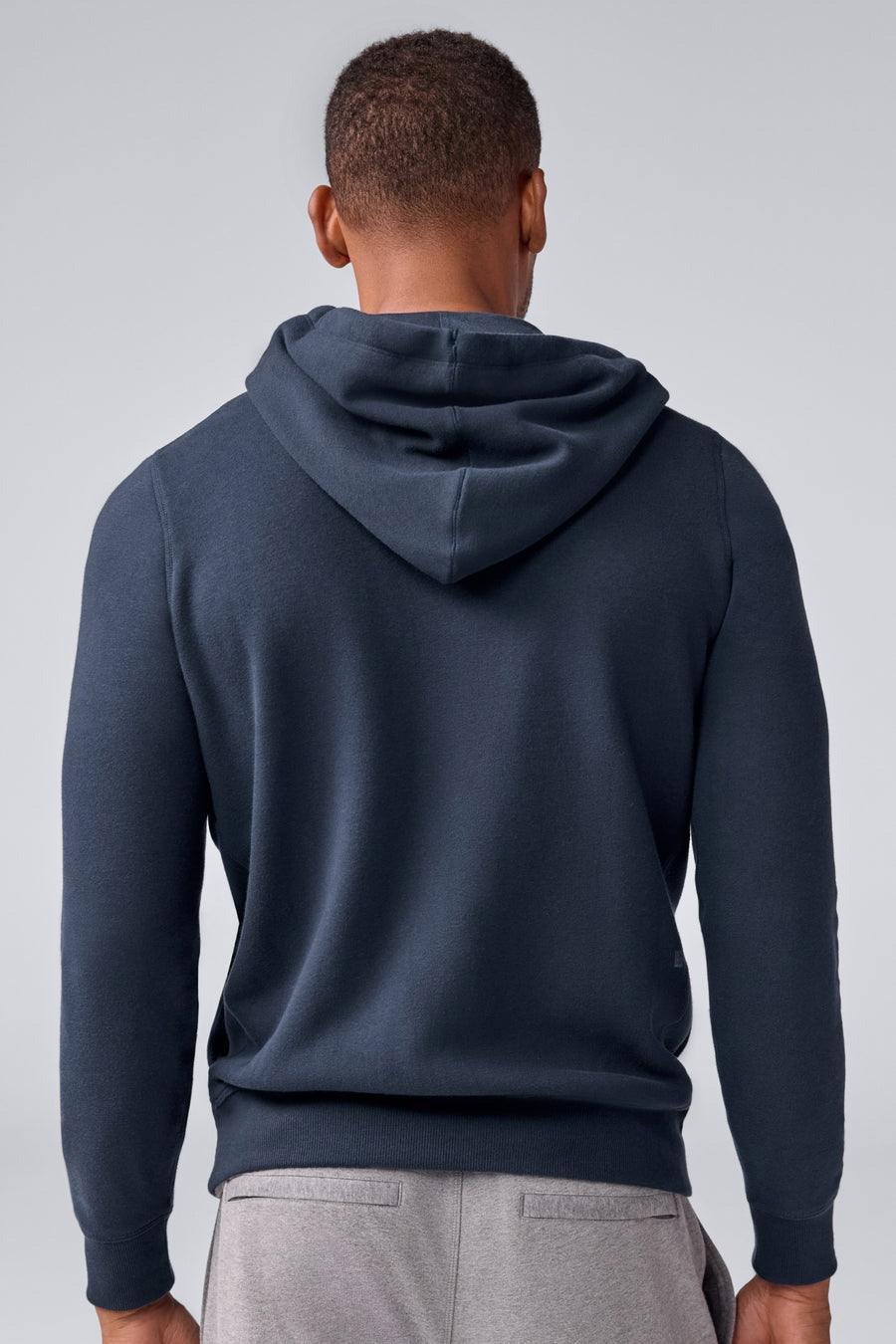 A man wears the MACK WELDON Ace Full-Zip Sweatshirt in True Navy (Bird's Eye) with light gray pants, standing against a plain light gray background and looking slightly to his left with a neutral expression.