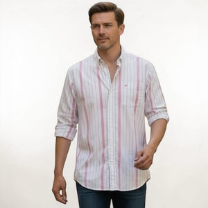 A man in a FRANK & EILEEN Finbar Men's Button Up Shirt in hot pink and blue stripe, blue jeans, and white sneakers walks confidently down a cobblestone street lined with stone buildings and potted plants on a sunny day.