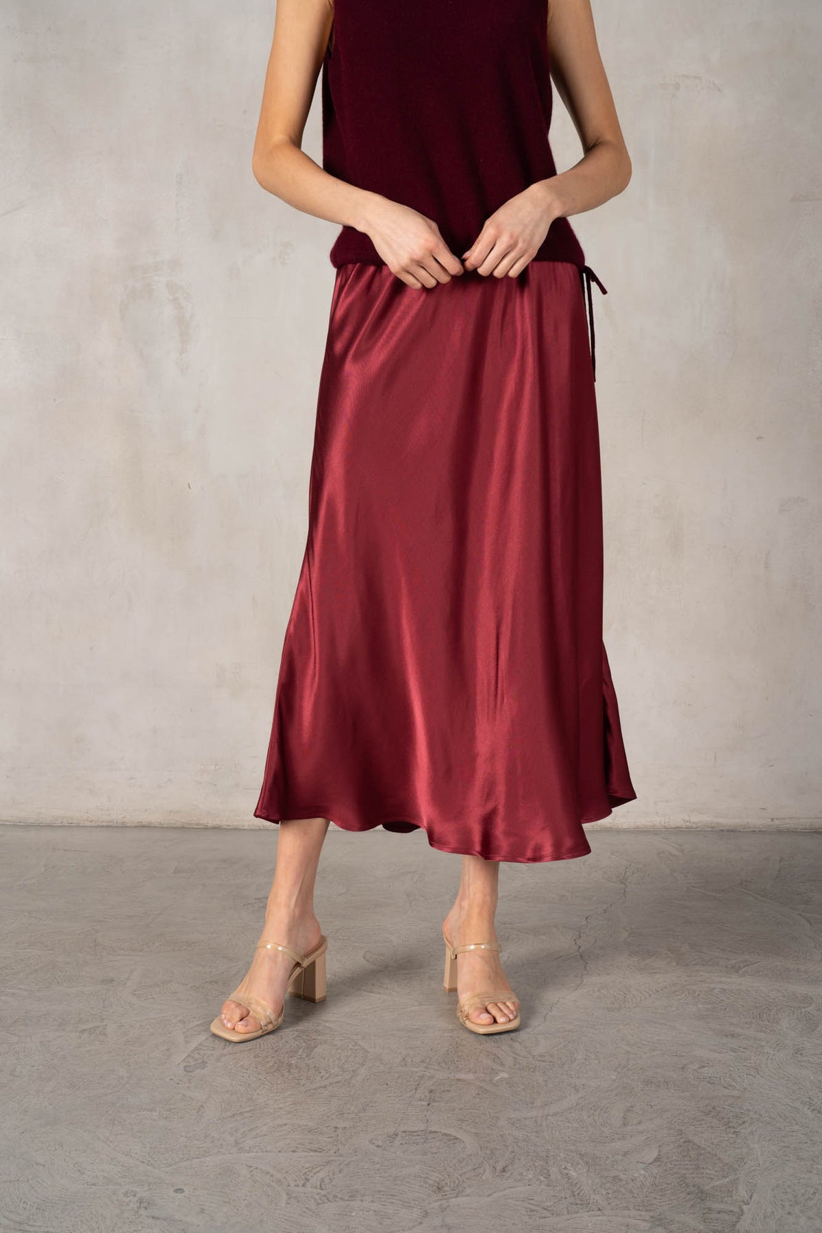 An older woman with long gray hair stands smiling in a dark red ROSE &amp; CROWN cardigan, the ROSE AND CROWN - SATIN MIDI SKIRT, and black knee-high boots against a plain white background.