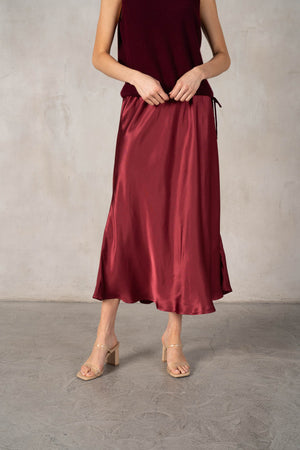 An older woman with long gray hair stands smiling in a dark red ROSE & CROWN cardigan, the ROSE AND CROWN - SATIN MIDI SKIRT, and black knee-high boots against a plain white background.