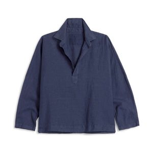 A woman with long brown hair stands outdoors among green plants and succulents, wearing the FRANK & EILEEN GABI Popover Shirt in Linen Navy and white pants.