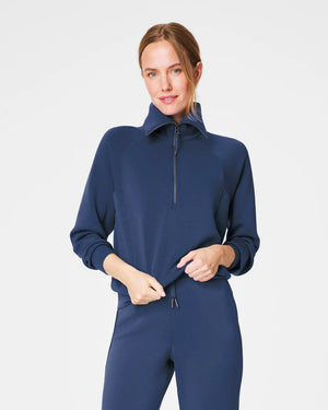 A person with short hair is wearing a SPANX AIRESSENTIALS HALF ZIP sweater in navy blue, made from lightweight spacer fabric, paired with black pants. Their arms are crossed over their chest as they stand against a plain, light grey background.