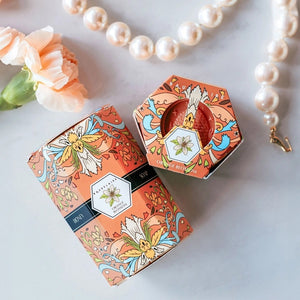 Hexagonal pink soaps with a honeycomb pattern are stacked beside a decorative orange and pink box labeled “BAUDELAIRE - ORANGE BLOSSOM HONEY SOAP 1.4 OZ QUARTET.” Each bar in this luxurious BAUDELAIRE set is triple-milled for rich lather and long-lasting Orange Blossom fragrance. The intricate floral designs on the box are complemented by a distinctive floral badge on top.