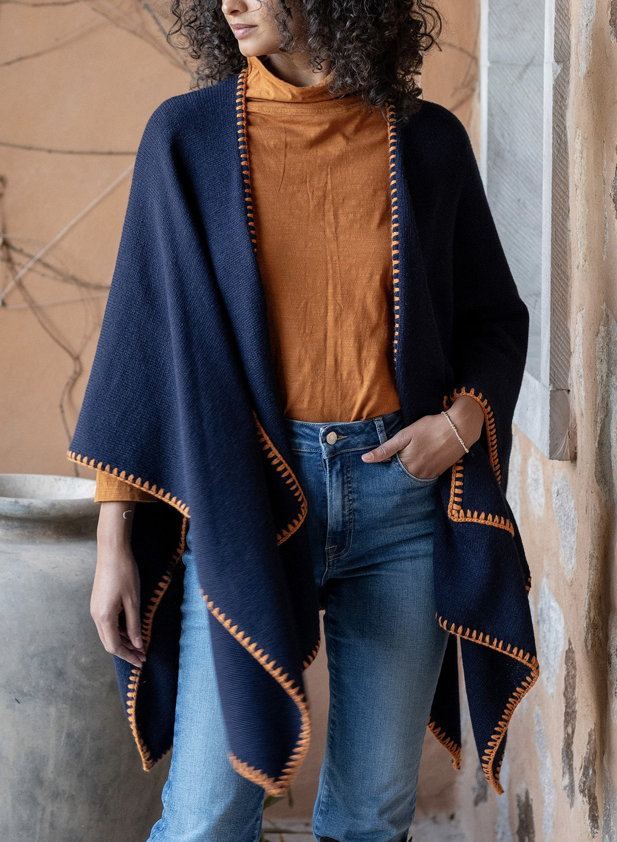 A person in a burnt orange turtleneck and blue jeans wears the MERSEA Whipstitch Travel Wrap in Navy/Golden, a lightweight wrap with gold trim, standing before a rustic wall and large vase.